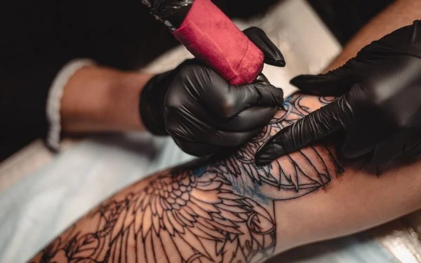 Eine nicht sichtbare Person mit schwarzen Schutzhandschuhen sticht mit einer Tätowiermaschine ein großes und aufwändiges Tattoo auf einem Bein.