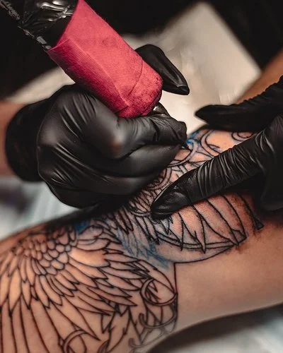 Eine nicht sichtbare Person mit schwarzen Schutzhandschuhen sticht mit einer Tätowiermaschine ein großes und aufwändiges Tattoo auf einem Bein.