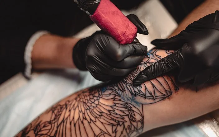 Eine nicht sichtbare Person mit schwarzen Schutzhandschuhen sticht mit einer Tätowiermaschine ein großes und aufwändiges Tattoo auf einem Bein.