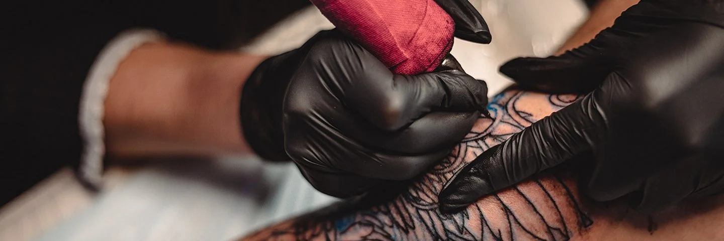 Eine nicht sichtbare Person mit schwarzen Schutzhandschuhen sticht mit einer Tätowiermaschine ein großes und aufwändiges Tattoo auf einem Bein.