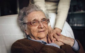 Eine ältere Frau mit Depressionen sitzt in einem Sessel, jemand hält ihre Hand.
