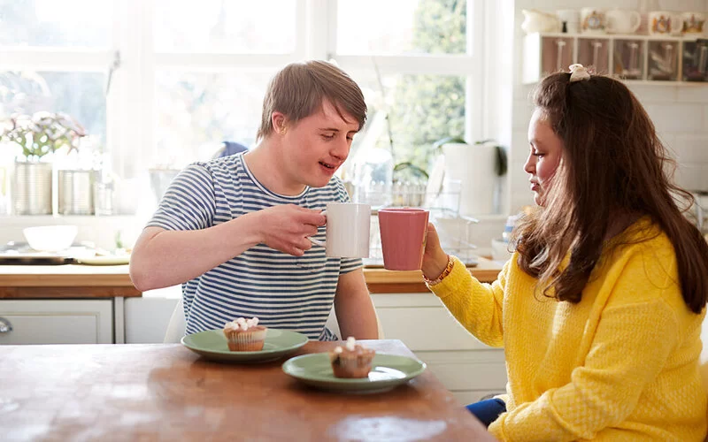Zwei Jugendliche, die in einer Wohnform für Menschen mit Behinderung leben, sitzen gemeinsam bei Kaffee und Kuchen.