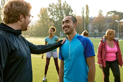 Zwei Männer stehen in sportlicher Kleidung auf einem Fußballfeld. Ein Mann legt seine Hand auf die Schulter des anderen Mannes. Im Hintergrund stehen zwei weitere Personen.