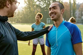 Zwei Männer stehen in sportlicher Kleidung auf einem Fußballfeld. Ein Mann legt seine Hand auf die Schulter des anderen Mannes. Im Hintergrund stehen zwei weitere Personen.
