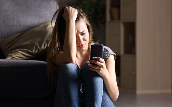 Eine Teenagerin sitzt traurig vor ihrem Bett mit dem Smartphone in der Hand.