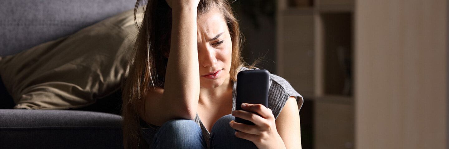 Eine Teenagerin sitzt traurig vor ihrem Bett mit dem Smartphone in der Hand.