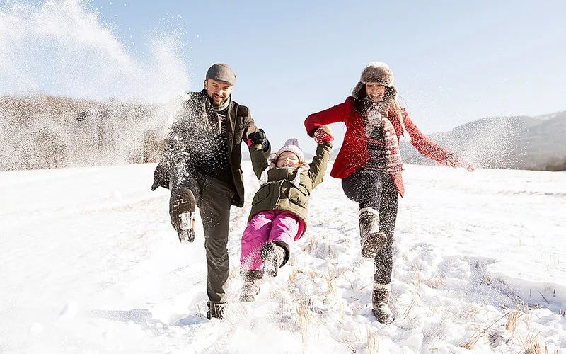 Ein Mann und eine Frau halten in ihrer Mitte ein Kind an den Händen und schwingen es über eine verschneite Wiese in die Luft. Dabei wirbelt Schnee auf. Alle Personen tragen warme Winterkleidung und lächeln.