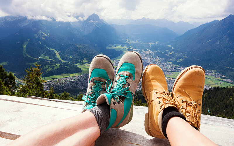 Zwei Wandernde legen ihre Unterschenkel mit Wanderstiefeln auf die Holzbrüstung eines Aussichtspunkts in den Alpen. Im Hintergrund liegt ein Gebirgstal.