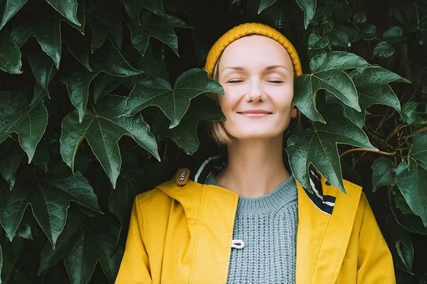 Ein Frau steht mit geschlossenen Augen in einem gelben Regenmantel vor einen Blätterwand.