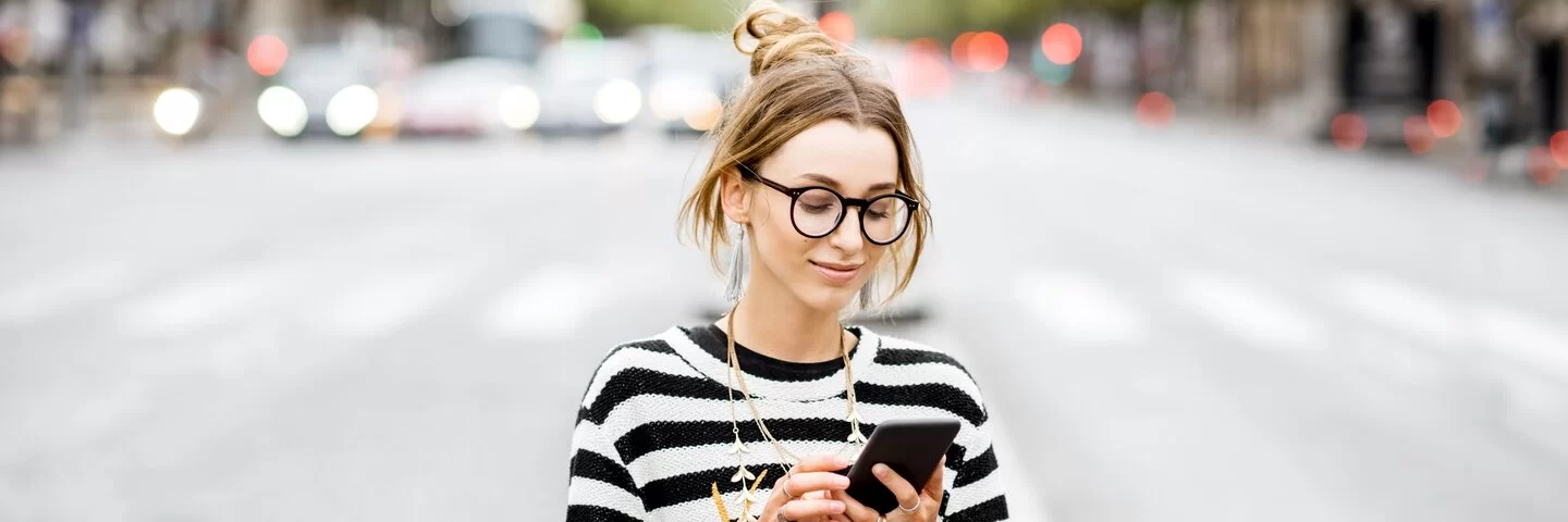 Junge Frau mit schwarzer Brille und Zebra-Pullover schaut auf ihr Handy.