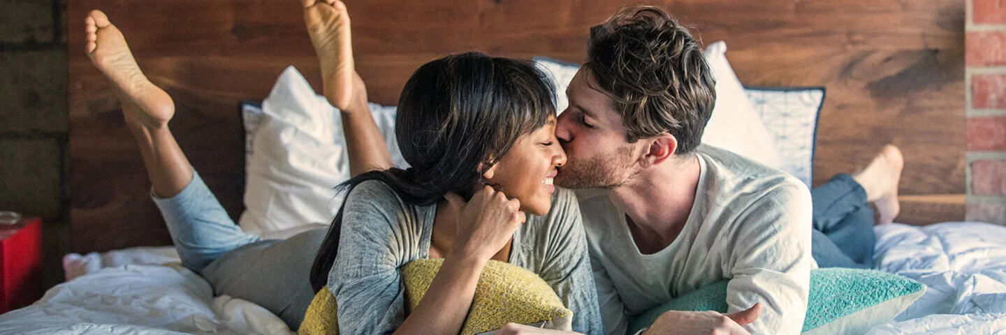 Ein Paar liegt glücklich lächelnd im Bett und teilt einen intimen Moment, bei dem der Mann die Frau auf die Wange küsst.