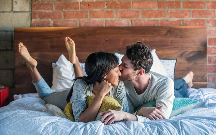 Ein Paar liegt glücklich lächelnd im Bett und teilt einen intimen Moment, bei dem der Mann die Frau auf die Wange küsst.