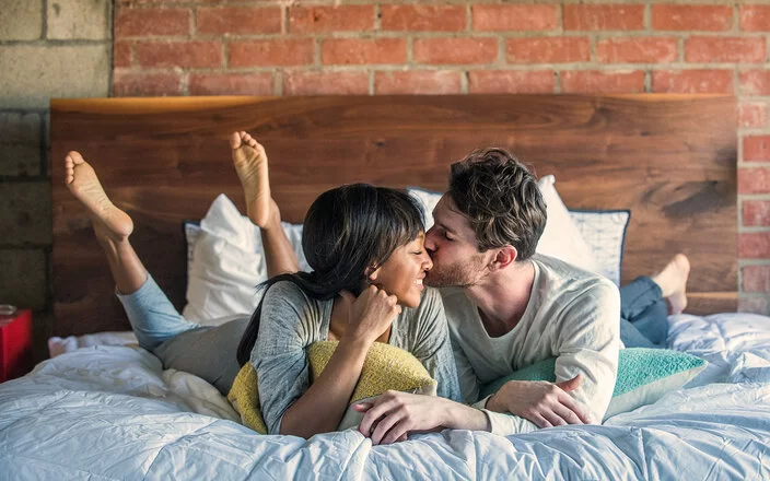 Ein Paar liegt glücklich lächelnd im Bett und teilt einen intimen Moment, bei dem der Mann die Frau auf die Wange küsst.