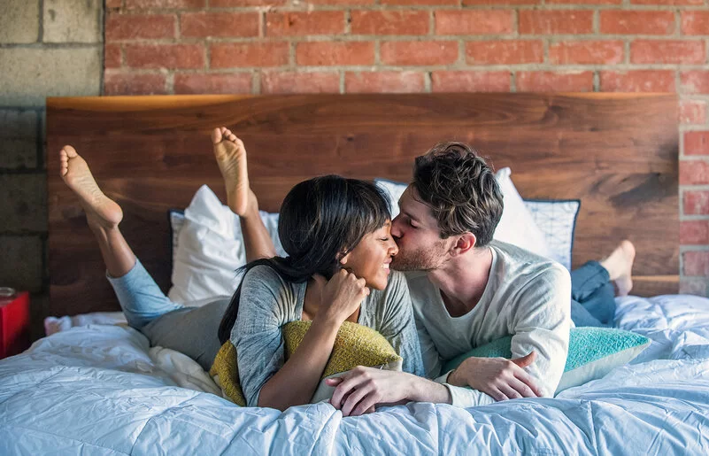 Ein Paar liegt glücklich lächelnd im Bett und teilt einen intimen Moment, bei dem der Mann die Frau auf die Wange küsst.