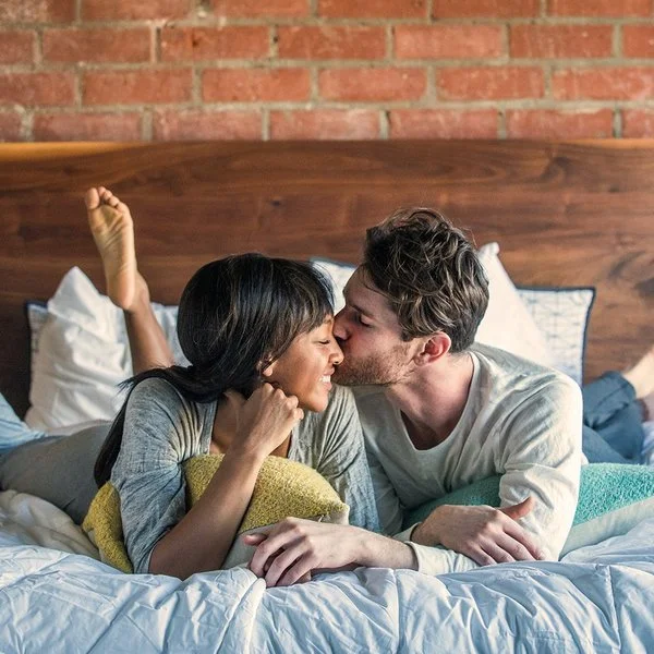Ein Paar liegt glücklich lächelnd im Bett und teilt einen intimen Moment, bei dem der Mann die Frau auf die Wange küsst.