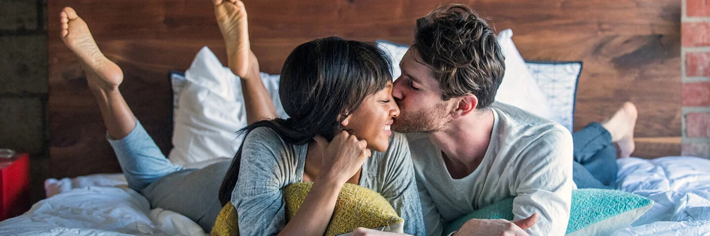 Ein Paar liegt glücklich lächelnd im Bett und teilt einen intimen Moment, bei dem der Mann die Frau auf die Wange küsst. 