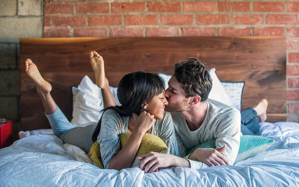 Ein Paar liegt glücklich lächelnd im Bett und teilt einen intimen Moment, bei dem der Mann die Frau auf die Wange küsst. 