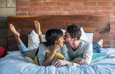 Ein Paar liegt glücklich lächelnd im Bett und teilt einen intimen Moment, bei dem der Mann die Frau auf die Wange küsst.