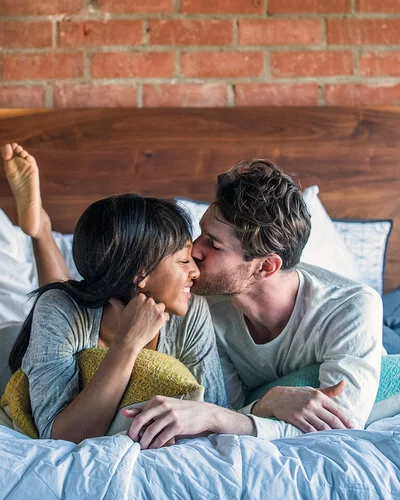 Ein Paar liegt glücklich lächelnd im Bett und teilt einen intimen Moment, bei dem der Mann die Frau auf die Wange küsst. 