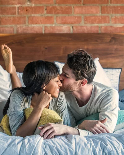 Ein Paar liegt glücklich lächelnd im Bett und teilt einen intimen Moment, bei dem der Mann die Frau auf die Wange küsst.