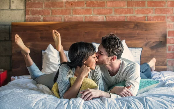 Ein Paar liegt glücklich lächelnd im Bett und teilt einen intimen Moment, bei dem der Mann die Frau auf die Wange küsst.