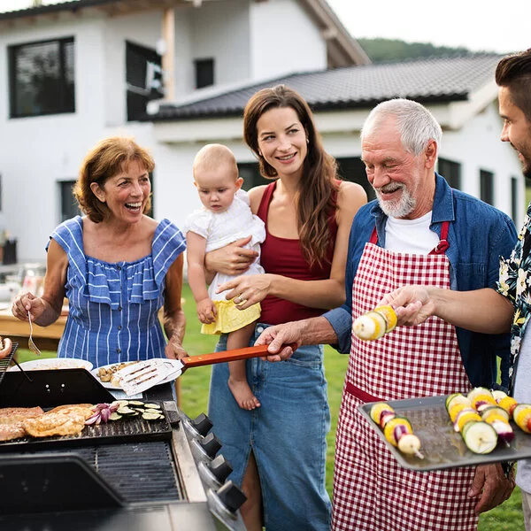 Eine Mehrgenerationenfamilie grillt im Garten umweltfreundlich mit Gas, Gemüse und Hähnchen.