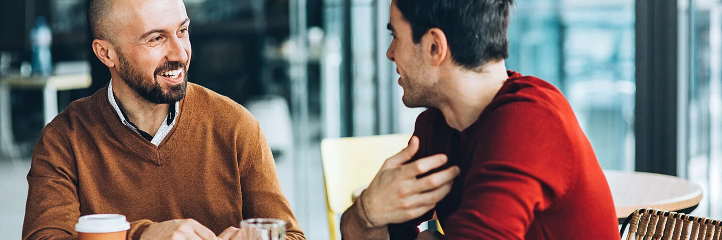 Zwei Männer sitzen am Tisch, einer erzählt etwas, der andere hört aktiv zu.