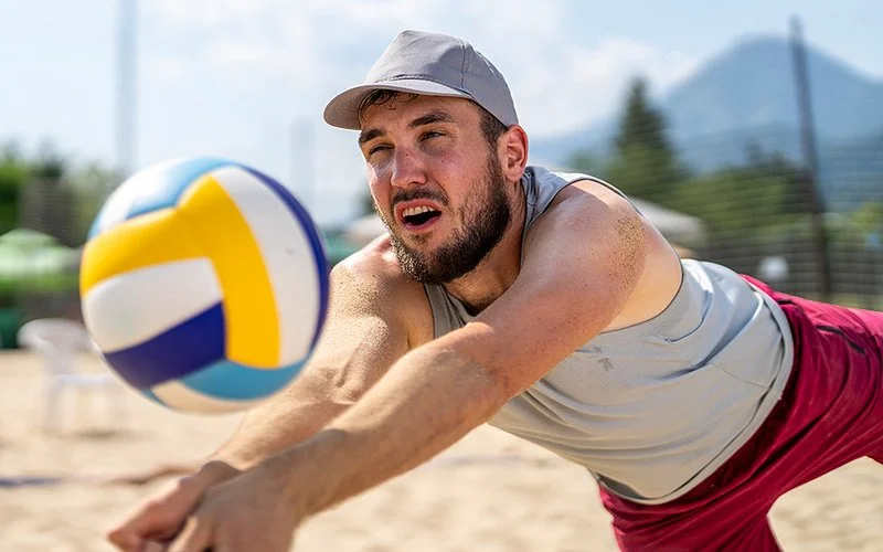 Ein sportlicher Mann in kurzer Sommerkleidung mit Kappe beim Beachvolleyball Spielen.