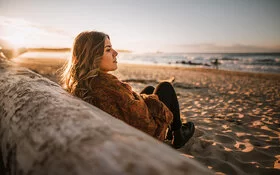 Frau sitzt an einen Stamm gelehnt am Strand und schaut auf das Meer.