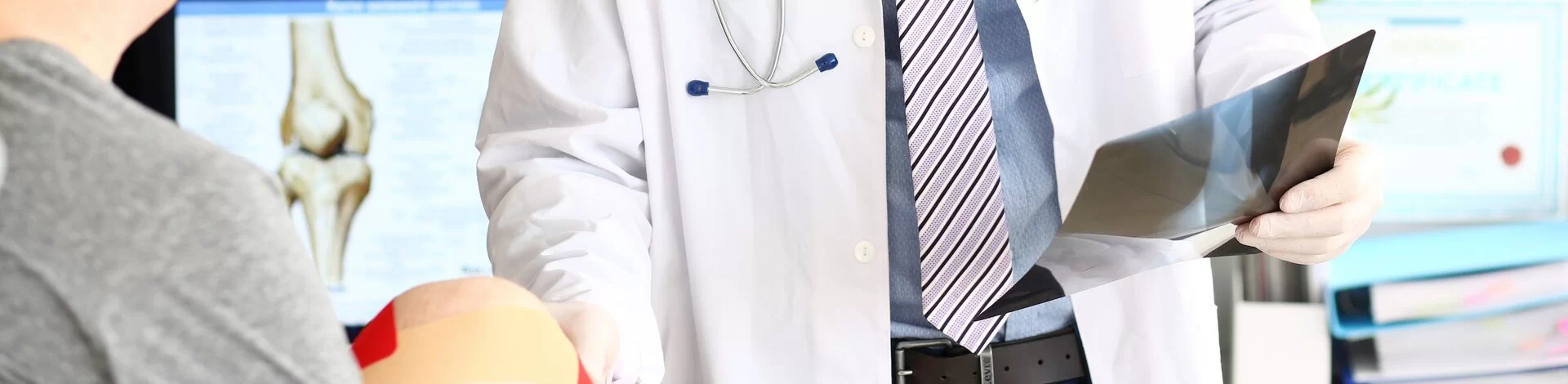 Arzt hält Röntgenaufnahme in der Hand. Patient sitzt mit bandagiertem Knie daneben.