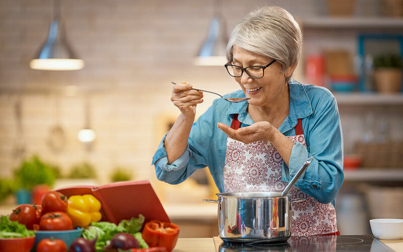 Ältere Dame bereitet Speisen in ihrer Küche zu und probiert vom Kochlöffel. Sie hat gelernt, intuitiv zu essen.