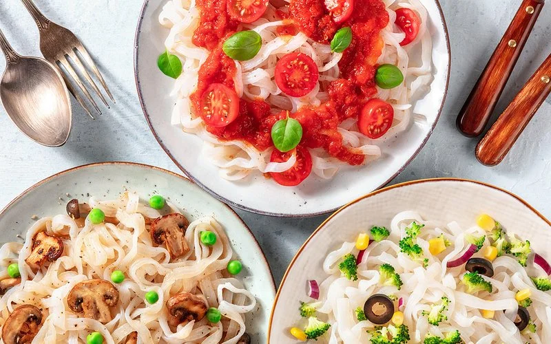 Drei Teller mit Konjaknudeln und unterschiedlichen Zutaten, wie Tomaten, Champignons und Brokkoli.