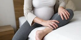 Eine schwangere Frau sitzt auf einem Bett. Ihr Gesicht ist nicht zu sehen. Das rechte Bein ist auf dem Boden aufgestellt, das linke liegt nach innen angewinkelt auf dem Bett. Mit den Händen massiert die Schwangere ihren linken Unterschenkel.