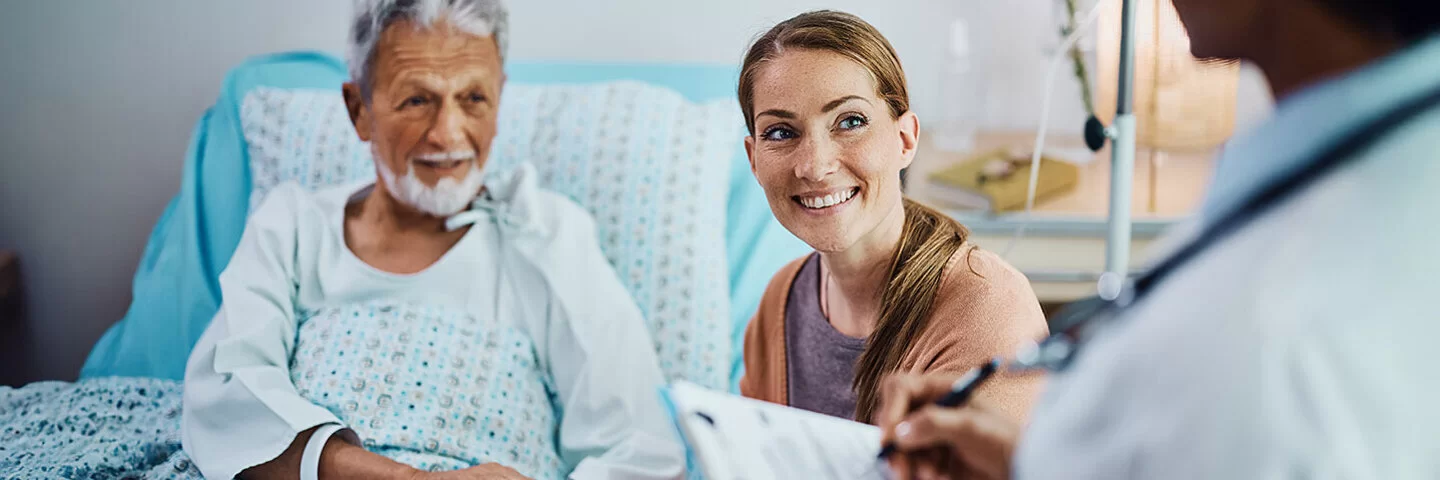 Ein älterer Patient liegt im Krankenhausbett und hält die Hand seiner Tochter. Die Tochter schaut zum Arzt.