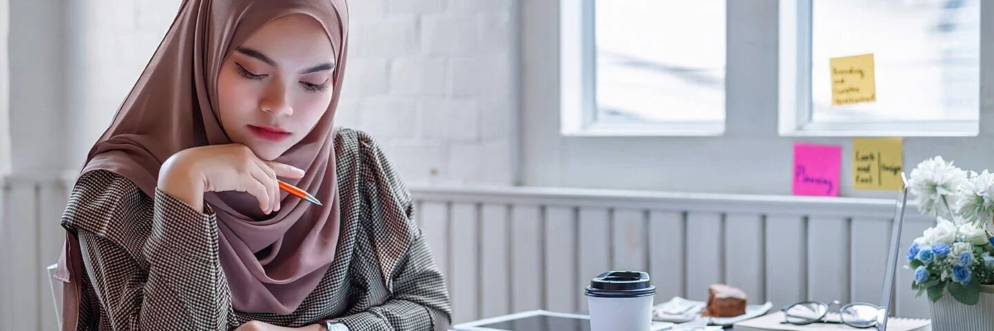 Eine Frau mit Kopftuch sitzt an einem Tisch und hält einen Bleistift in der rechten Hand, während sie auf ein paar Arbeitsblätter schaut.