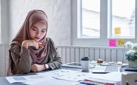 Eine Frau mit Kopftuch sitzt an einem Tisch und hält einen Bleistift in der rechten Hand, während sie auf ein paar Arbeitsblätter schaut.