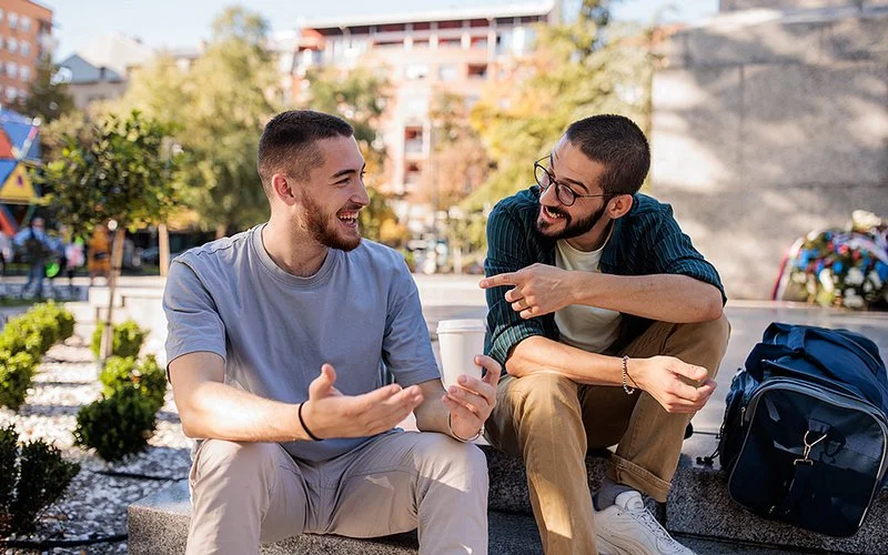 Zwei junge Männer sitzten an einem sonnigen Tag draußen in einer öffentlichen Grünanlage und unterhalten sich lachend.
