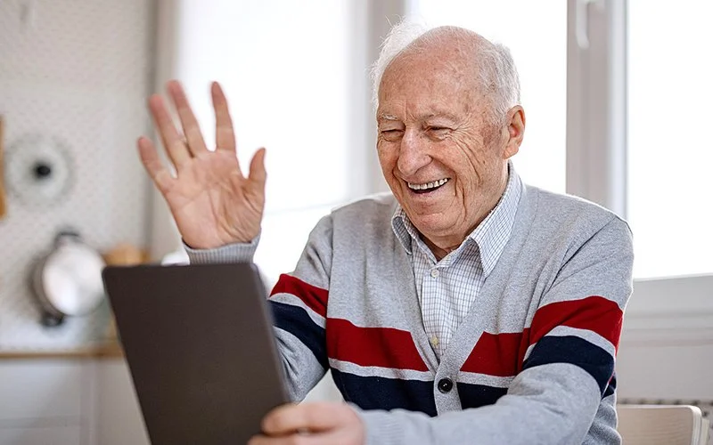 Am Küchentisch sitzt ein alter Mann. In seiner linken Hand hält er ein Tablet. Er blickt auf das Display, lacht und winkt dem Bildschirm mit der rechten Hand zu.