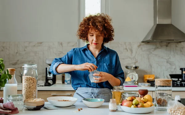Eine jüngere Frau mit blauer Bluse steht in der Küche und bereitet ein Müsli zu, unter anderem mit Vitamin B1-haltigen Haferflocken, Milch und Obst.