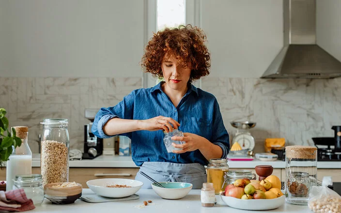 Eine jüngere Frau mit blauer Bluse steht in der Küche und bereitet ein Müsli zu, unter anderem mit Vitamin B1-haltigen Haferflocken, Milch und Obst.