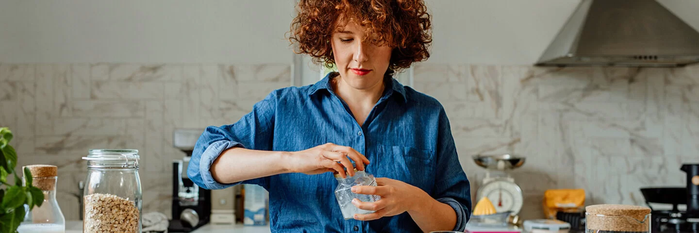 Eine jüngere Frau mit blauer Bluse steht in der Küche und bereitet ein Müsli zu, unter anderem mit Vitamin B1-haltigen Haferflocken, Milch und Obst.