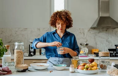 Eine jüngere Frau mit blauer Bluse steht in der Küche und bereitet ein Müsli zu, unter anderem mit Vitamin B1-haltigen Haferflocken, Milch und Obst.
