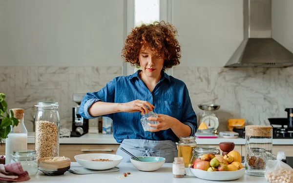 Eine jüngere Frau mit blauer Bluse steht in der Küche und bereitet ein Müsli zu, unter anderem mit Vitamin B1-haltigen Haferflocken, Milch und Obst.