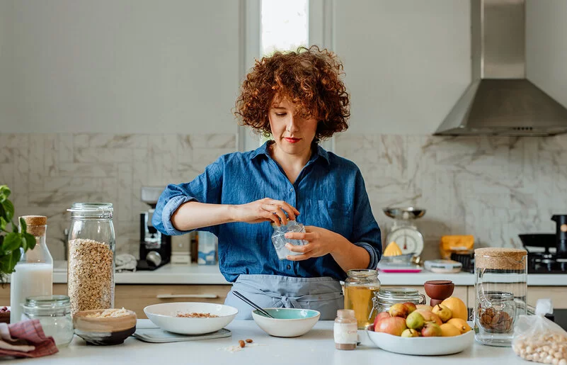 Eine jüngere Frau mit blauer Bluse steht in der Küche und bereitet ein Müsli zu, unter anderem mit Vitamin B1-haltigen Haferflocken, Milch und Obst.