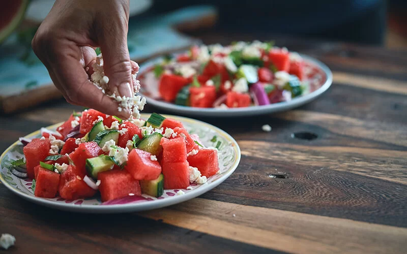 Eine Person krümmelt Fetakäse auf einen gesunden Salat aus Wassermelone und Gurke.
