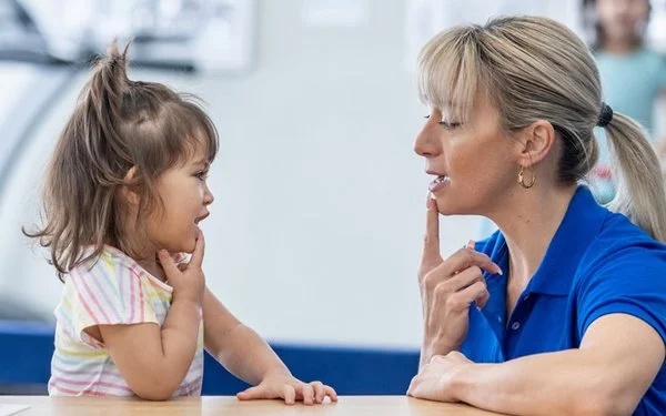 Therapeutin sitzt einem kleinen Kind gegenüber und zeigt mit dem Finger auf den Mund; beide üben in einem hellen Therapieraum.