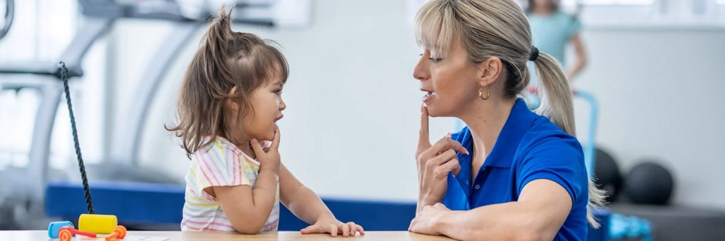 Therapeutin sitzt einem kleinen Kind gegenüber und zeigt mit dem Finger auf den Mund; beide üben in einem hellen Therapieraum.