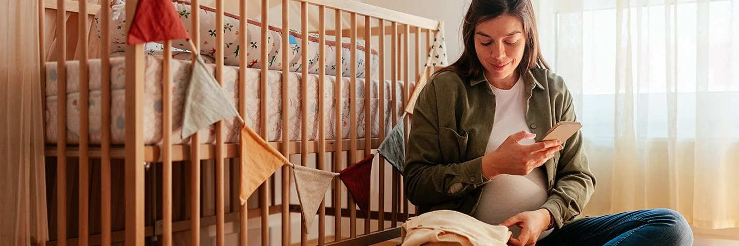 Eine schwangere Frau sitzt auf einem bunten Teppichboden eines gemütlich eingerichteten Kinderzimmers. Neben ihr steht ein Kinderbett mit einer bunten Wimpelkette. Sie blickt auf ein Smartphone, dass sie in der Hand hält.