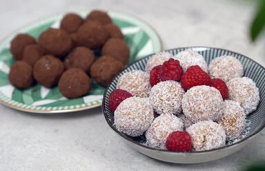 Im Vordergrund Kokos-Himbeer Energy Balls nach einem Rezept von Felicitas Then, angerichtet auf einem schwarz-weiß gestreiften Teller, garniert mit Himbeeren. Im Hintergrund Schoko-Erdnuss Energy Balls auf einem grün-weißem Teller.