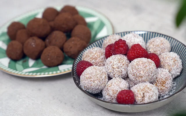 Im Vordergrund Kokos-Himbeer Energy Balls nach einem Rezept von Felicitas Then, angerichtet auf einem schwarz-weiß gestreiften Teller, garniert mit Himbeeren. Im Hintergrund Schoko-Erdnuss Energy Balls auf einem grün-weißem Teller.